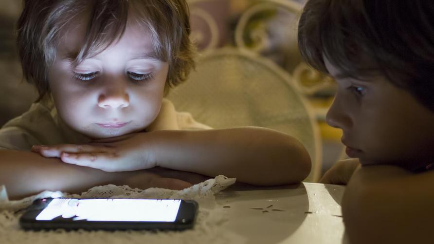 Alltag mit Kindern: Two young boys are focused on a smartphone illuminated in front of them while sitting at a table in a cozy café during the evening, surrounded by soft lighting and a relaxed atmosphere.