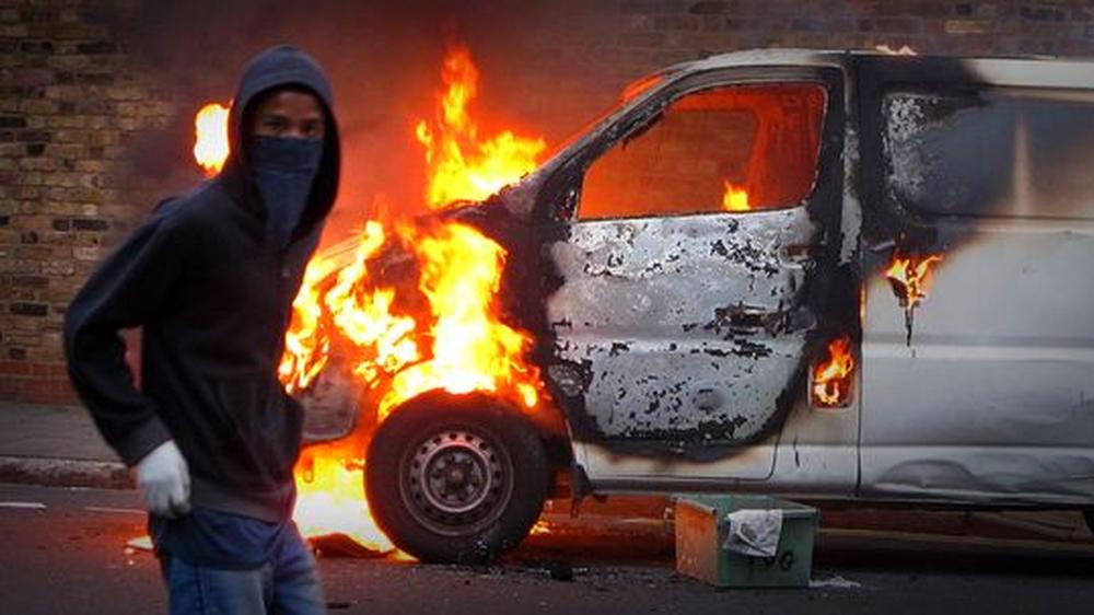 Krawalle in London: Ein vermummter Jugendlicher steht vor einem brennenden Auto.