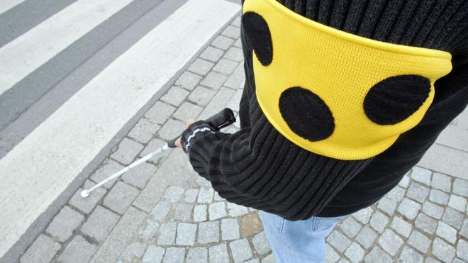 Soziales: Ein blinder Mann geht mit seinem weißen Taststock und einer Armbinde mit drei Punkten über eine Straße in Hannover.