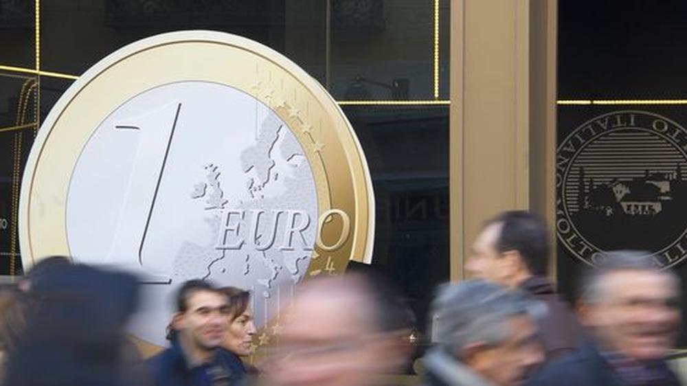 Menschen laufen in Madrid an einem Schaufenster mit einem großen Euro-Aufkleber vorbei.