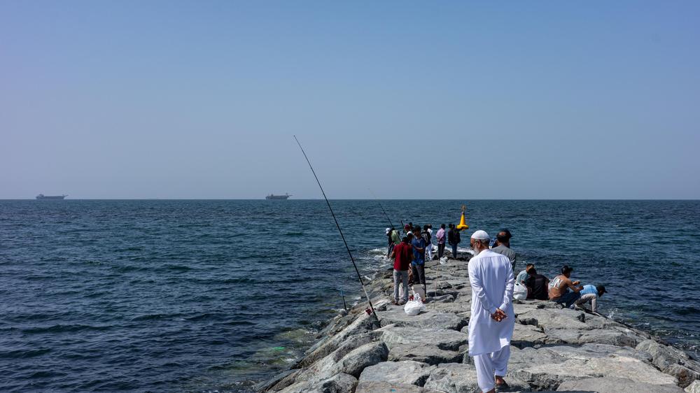  Menschen stehen am 22. März auf einem Wellenbrecher, im Hintergrund liegen Handelsschiffe nahe der Straße von Hormus vor Anker.