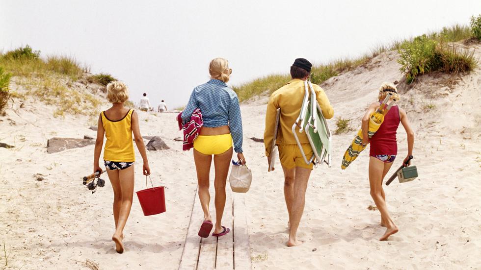 Urlaub mit den Eltern: 1970s FAMILY, MOTHER, FATHER, TWO BOYS WALKING ON TO BEACH CARRYING CHAIRS UMBRELLA GEAR.